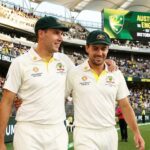 Australian cricketers Scott Boland and Brendan Doggett walk side-by-side on a stadium field, wearing baggy green caps and white Test uniforms. Boland has his arm around Doggett's shoulder, and both are smiling. The background shows a cheering crowd and a scoreboard reading "AUSTRALIA VS ENGLAND."