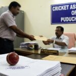 A photo inside an office with a sign for the "CRICKET ASSOCIATION OF PUDUCHERRY". A man is handing a large envelope to another official across a desk. In the foreground, there is a stack of documents stamped "DOMICILE CERTIFICATE - PUDUCHERRY" next to a red cricket ball.