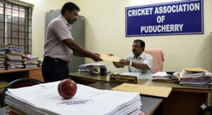 A photo inside an office with a sign for the "CRICKET ASSOCIATION OF PUDUCHERRY". A man is handing a large envelope to another official across a desk. In the foreground, there is a stack of documents stamped "DOMICILE CERTIFICATE - PUDUCHERRY" next to a red cricket ball.