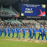 The Italian national cricket team, wearing blue jerseys with "ITALIA" across the chest, walks in a line onto the green field at Eden Gardens stadium at night. A large digital scoreboard in the background reads "ICC MEN'S T20 WORLD CUP 2026: ITALY VS SCOTLAND" and "ITALY DEBUT" in bold letters. Spectators are visible in the stands.