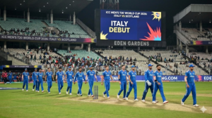 The Italian national cricket team, wearing blue jerseys with "ITALIA" across the chest, walks in a line onto the green field at Eden Gardens stadium at night. A large digital scoreboard in the background reads "ICC MEN'S T20 WORLD CUP 2026: ITALY VS SCOTLAND" and "ITALY DEBUT" in bold letters. Spectators are visible in the stands.